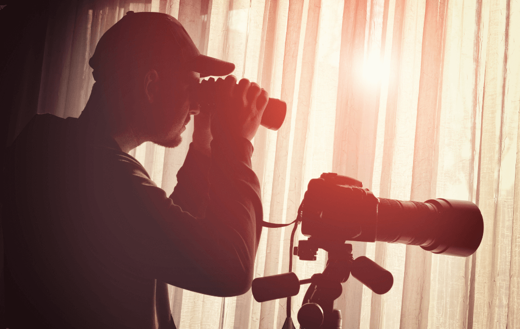 Man looking out window with binoculars next to camera with large lens